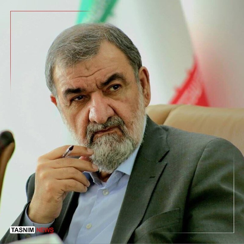 A bearded man in a gray suit holds a pen near his face, seated before a green, white, and red flag, with a "TASNIM NEWS" logo visible.
