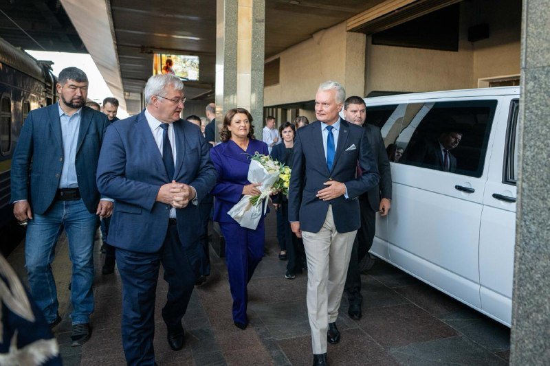 A group of formally dressed individuals, including a woman holding a bouquet of white flowers, walk through a covered arrival area beside a white van.