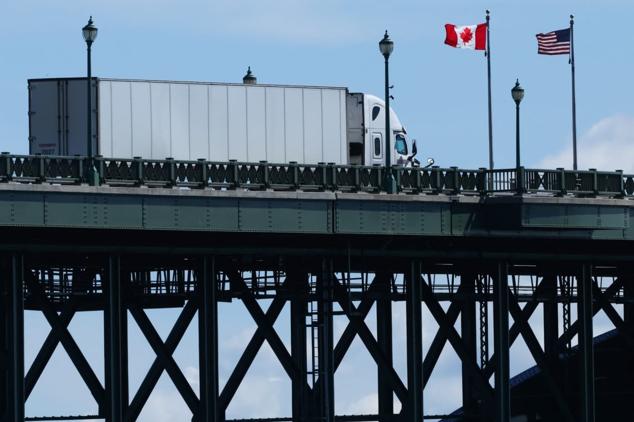 A white semi-truck with a trailer crosses a green steel bridge, passing beneath Canadian and American flags flying on flagpoles against a blue sky.