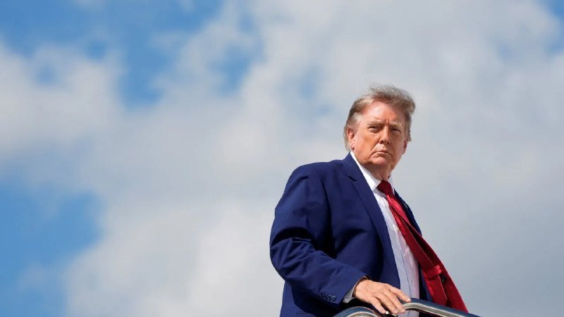 An older man with light hair, wearing a dark blue suit and red tie, stands outdoors against a partly cloudy sky, looking toward the camera.