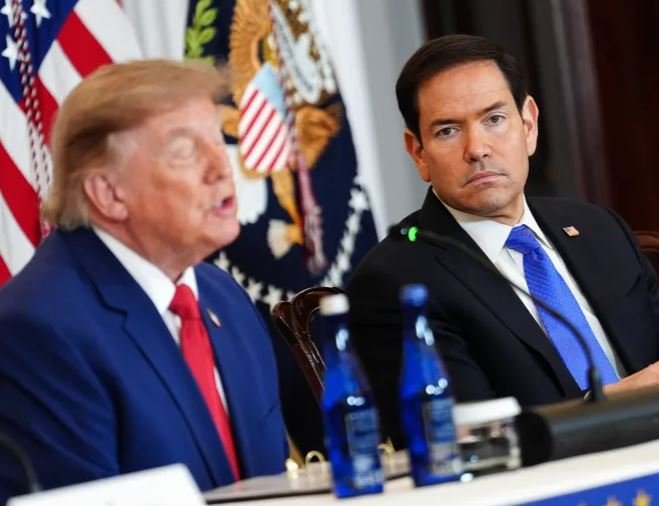 Two men in suits sit at a table with water bottles, an American flag and a presidential seal flag visible behind them.