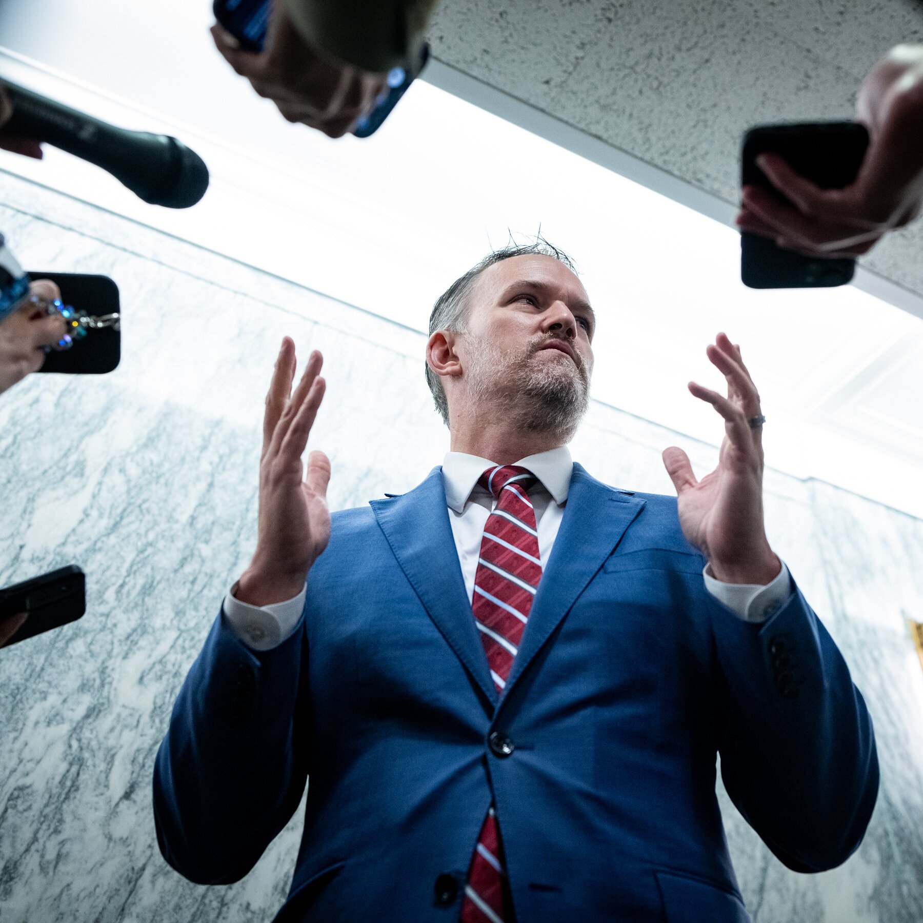 A bearded man in a blue suit and red striped tie gestures with both hands raised while speaking to surrounding microphones and recording devices.