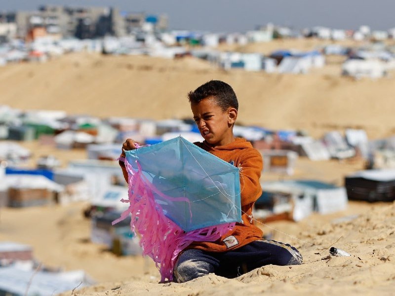 A young boy in an orange hoodie sits on sand, holding a blue kite with pink streamers, with a sprawling encampment of makeshift shelters visible in the background.