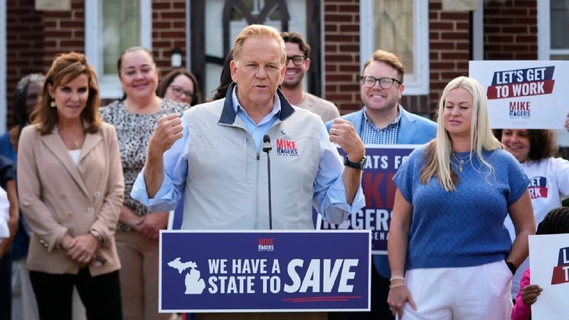 A man wearing a "MIKE ROGERS" vest speaks at a podium with a "WE HAVE A STATE TO SAVE" sign, surrounded by supporters holding "LET'S GET TO WORK" signs.