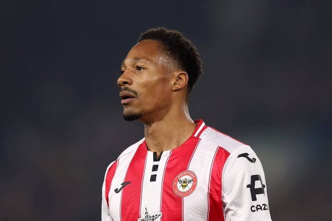 A soccer player wearing a red and white striped jersey with a club crest and sponsor logos stands on a pitch, looking off to the side during a match.