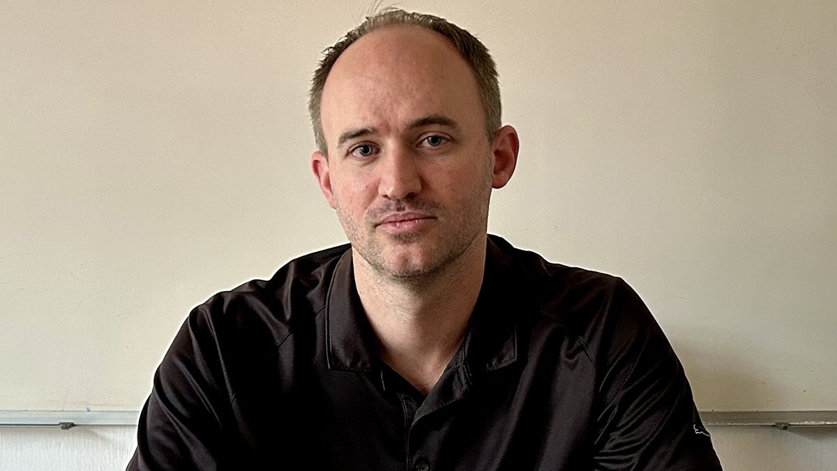 A man with short brown hair and a light beard, wearing a black collared shirt, looks directly at the camera against a plain off-white wall.