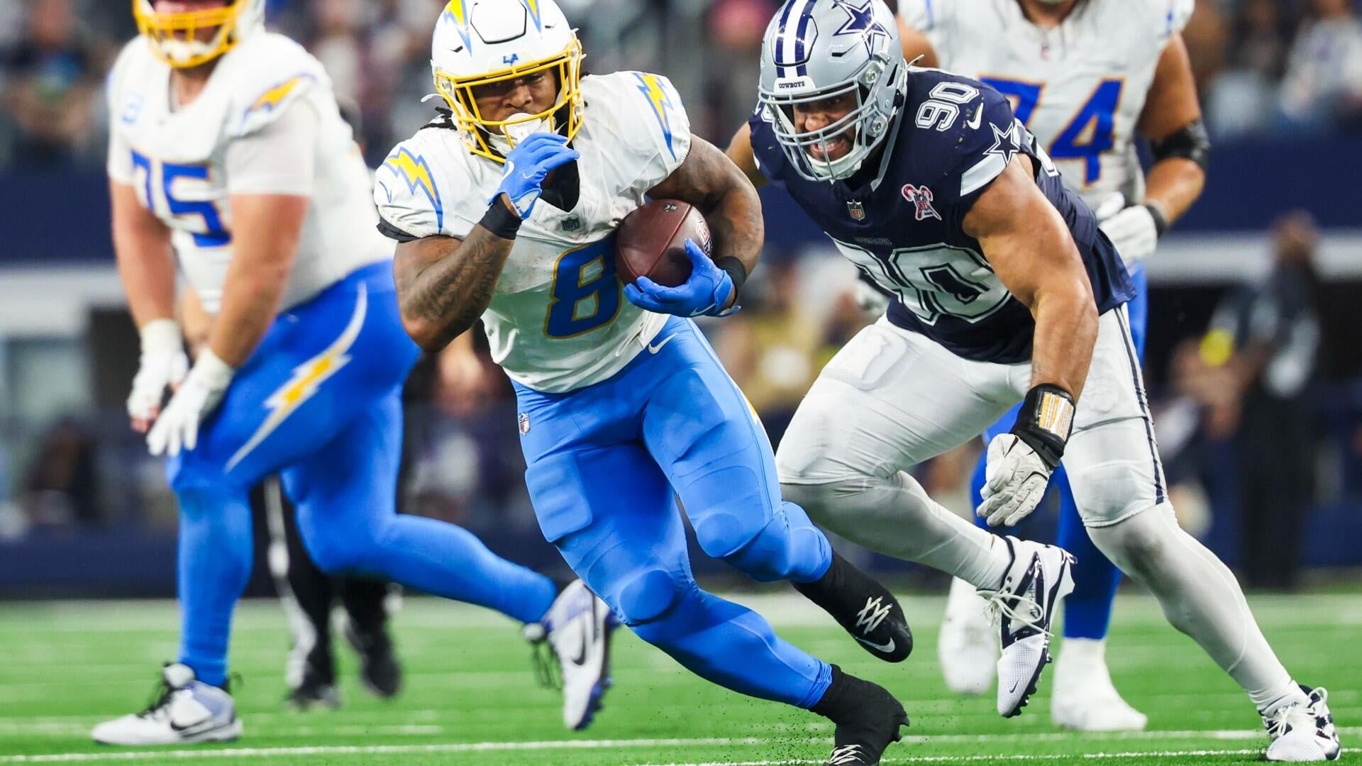 A Chargers player in a white and blue uniform carries the football while being pursued by a Cowboys defender wearing number 98 in a navy jersey.