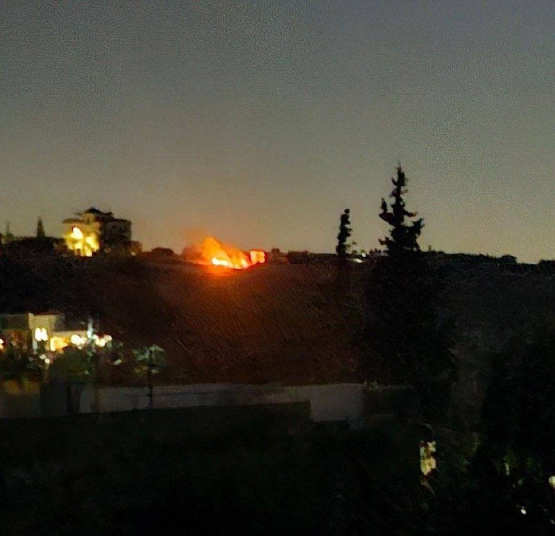 A bright orange fire burns on a hillside at dusk, with a lit building and silhouetted trees visible nearby.