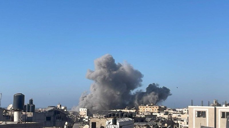 Large plumes of gray smoke rise above densely packed low-rise buildings under a clear blue sky.
