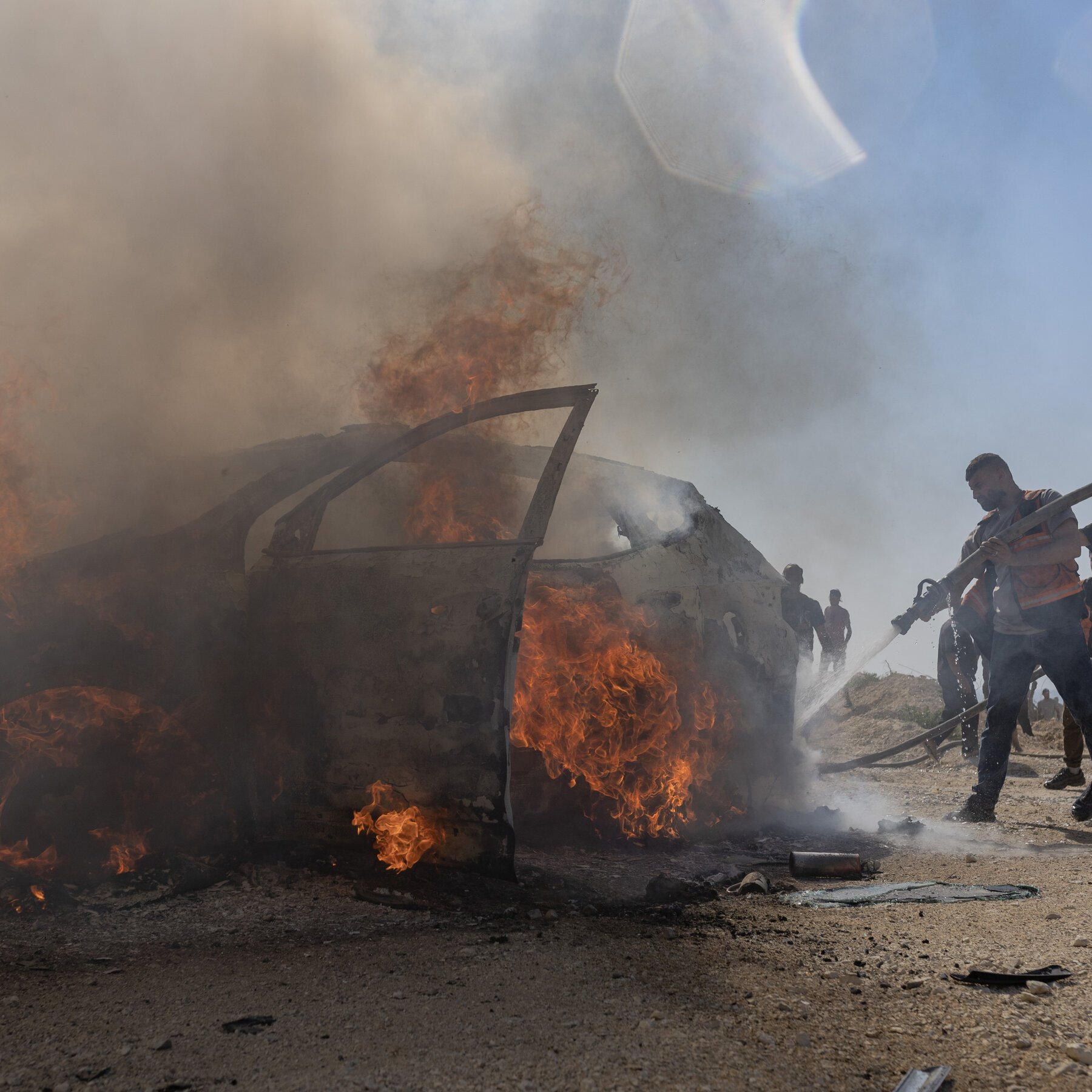 Men in high-visibility vests spray water from a hose onto a burning, smoke-engulfed vehicle on a dirt surface.