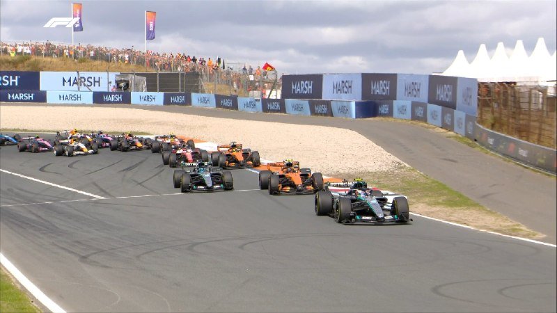 A pack of Formula 1 cars races down a track, led by a silver Mercedes, with "MARSH" branded barriers and a crowd of spectators visible in the background.