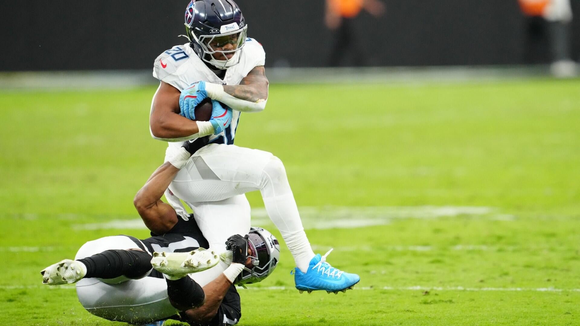 A football player in a white Tennessee Titans jersey carrying the ball is tackled to the ground by an opponent in a black and silver uniform on a green field.