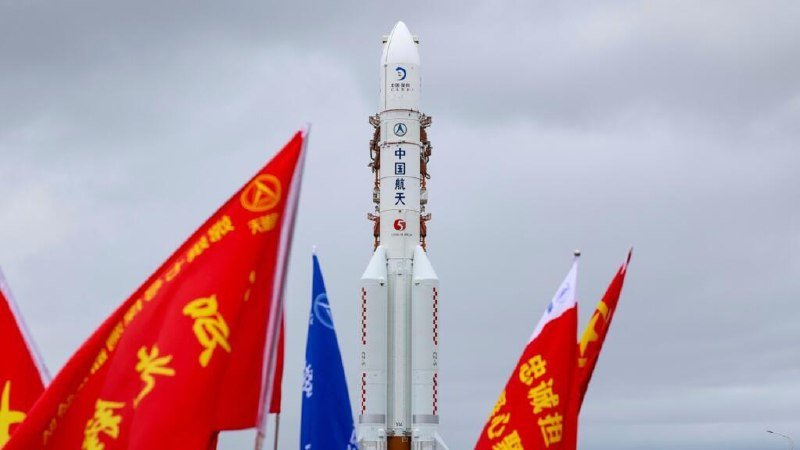 A white rocket with Chinese characters and logos stands vertically on a launch pad, flanked by red, blue, and multicolored flags under an overcast sky.