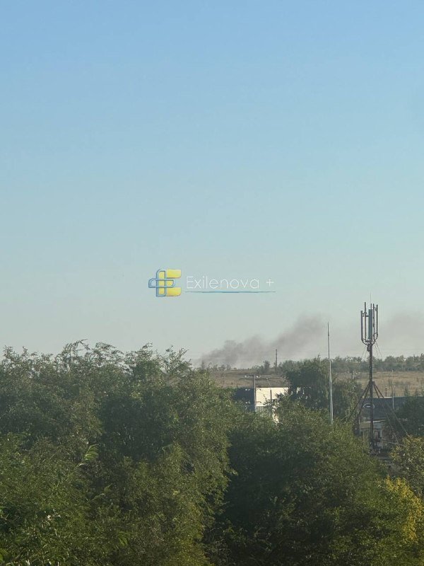 Dark smoke rises in the distance behind green foliage, a communications tower, and a building under a clear blue sky.