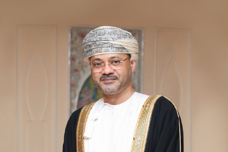 A bearded man wearing glasses, a patterned turban, and traditional Arab attire poses indoors in front of an ornately decorated wall with floral tilework.
