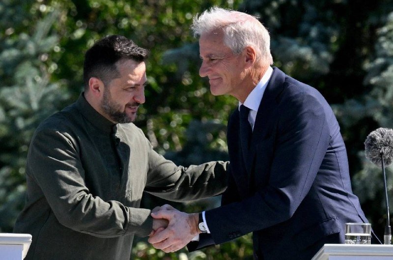 Two men in dark clothing shake hands and smile at each other outdoors, with a microphone visible on the right and green foliage in the background.