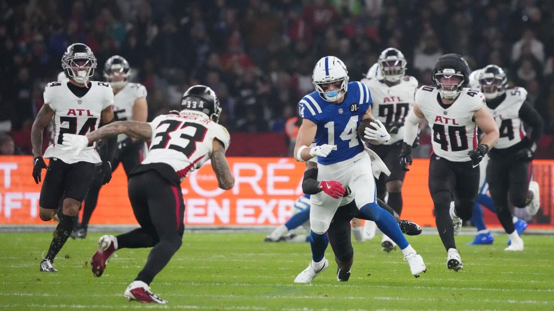 A football player in a blue #14 jersey carries the ball while being pursued by several players in white "ATL" jerseys during a game in a stadium.