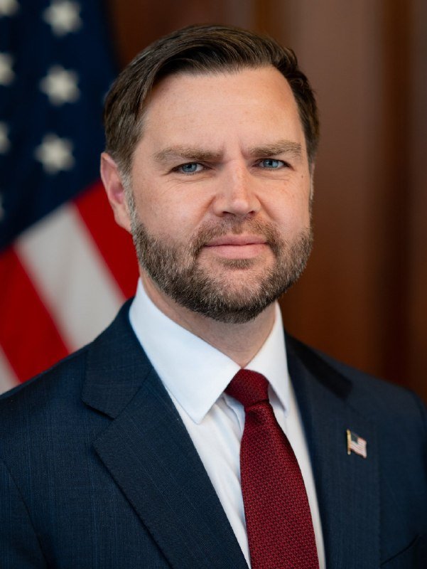 Official portrait of a bearded man in a dark suit, white shirt, and burgundy tie with an American flag pin, standing before a U.S. flag.