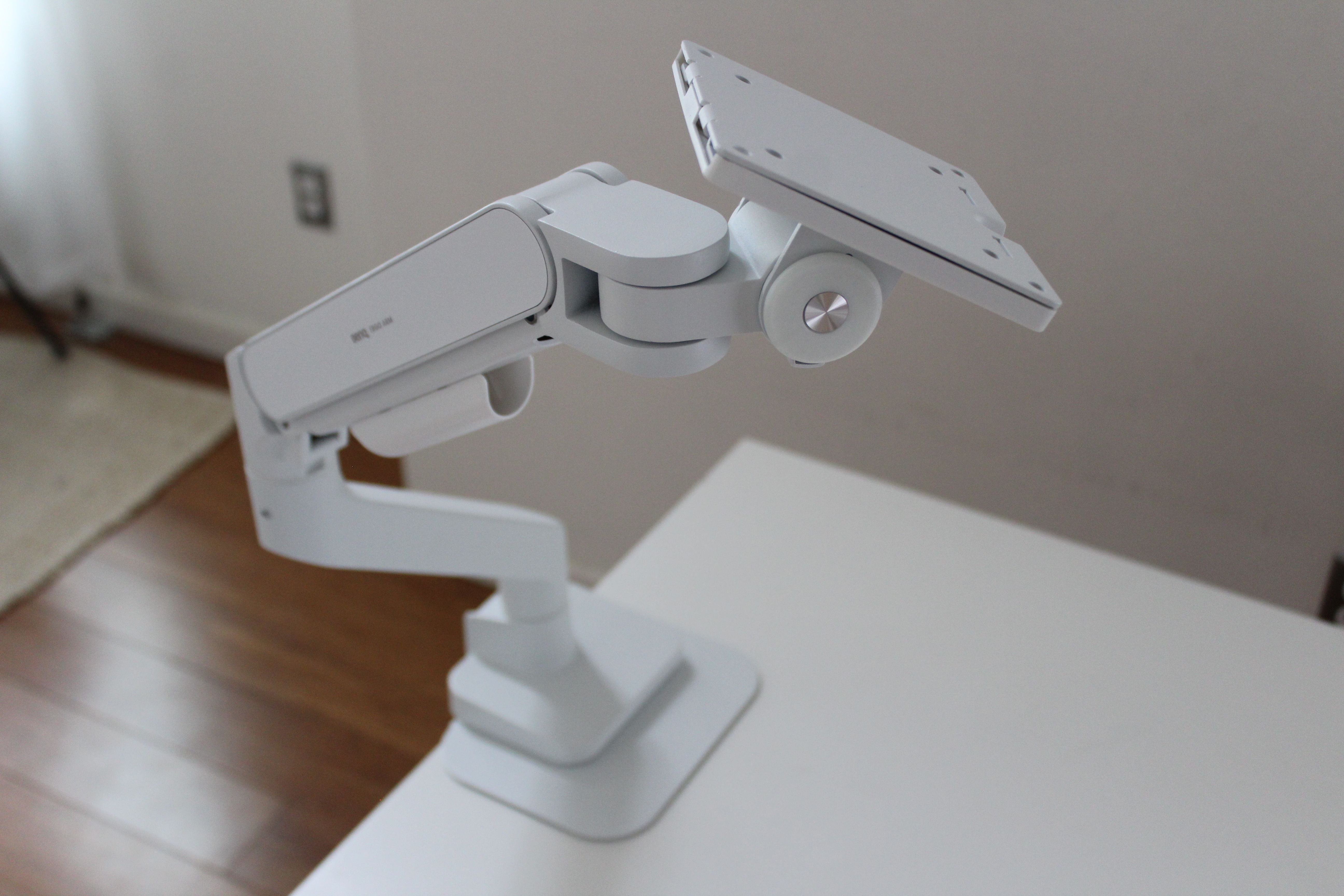 A white articulated mounting arm with an adjustable mounting plate at one end and a weighted base sits on a white table.