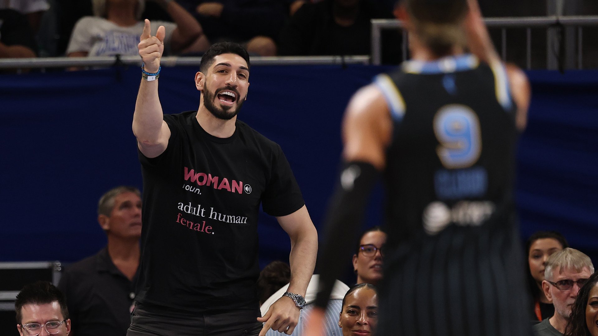 A smiling, bearded man in a "WOMAN: adult human female" t-shirt gestures with a raised finger while standing courtside near a player in a black number 9 jersey.