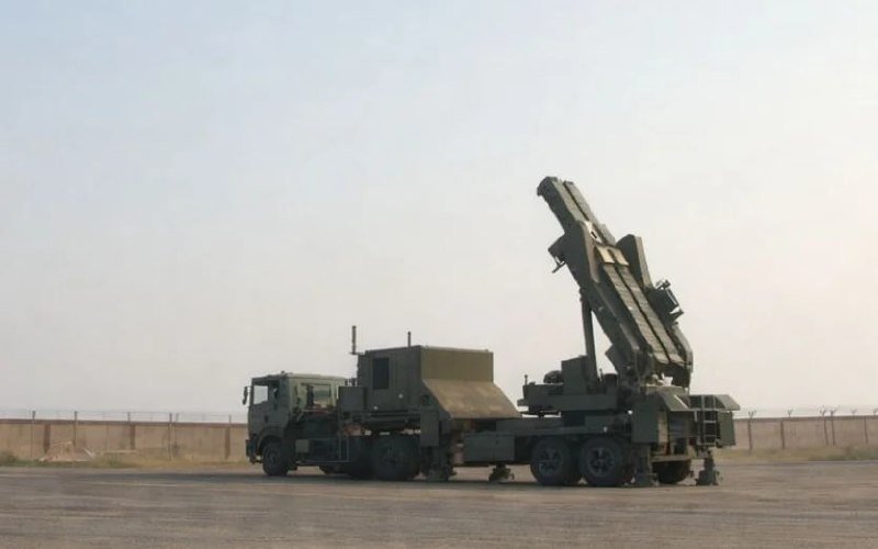 A military truck carrying a raised, angled missile launcher system is parked on an open tarmac behind a perimeter fence.