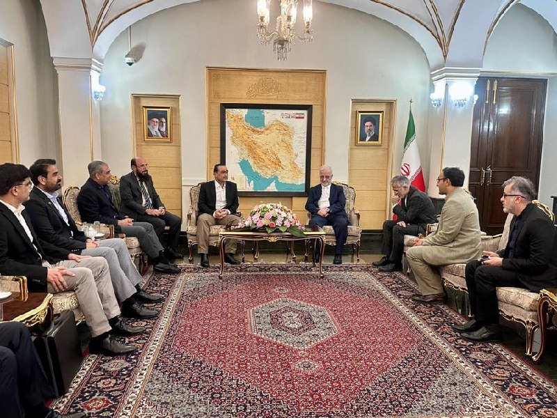 Two groups of men in suits sit facing each other on ornate sofas in a formal room featuring a large map of Iran, an Iranian flag, chandeliers, and framed portraits on the walls.