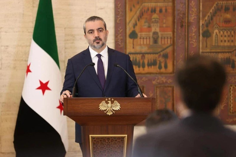A bearded man in a dark suit stands at a wooden podium bearing a golden eagle emblem, speaking into microphones beside a flag with green, white, and black stripes and red stars.