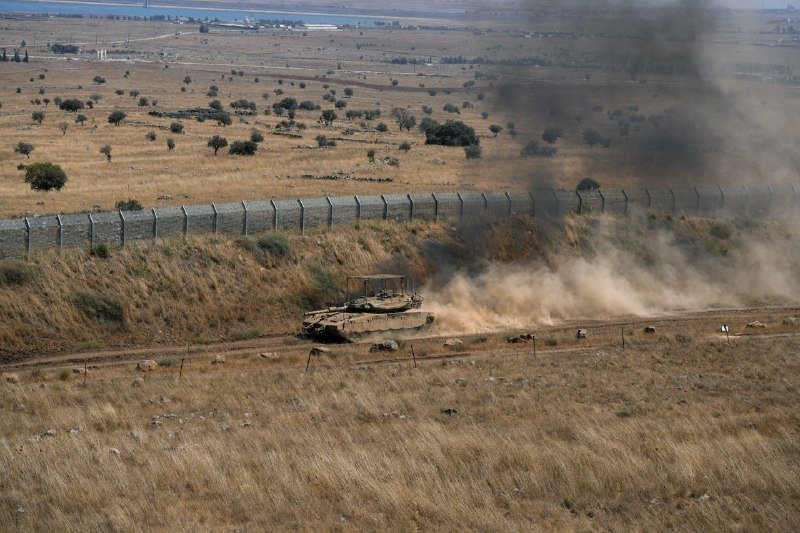 A tan military bulldozer kicks up a large dust cloud while operating along a dirt track beside a tall, barbed-wire border fence in a dry, grassy landscape.