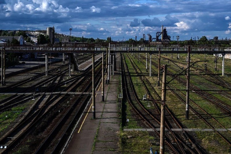 Multiple railway tracks extend through an industrial rail yard beneath a partly cloudy sky, with overhead electrical lines, a pedestrian overpass, and distant factory structures visible on the horizon.