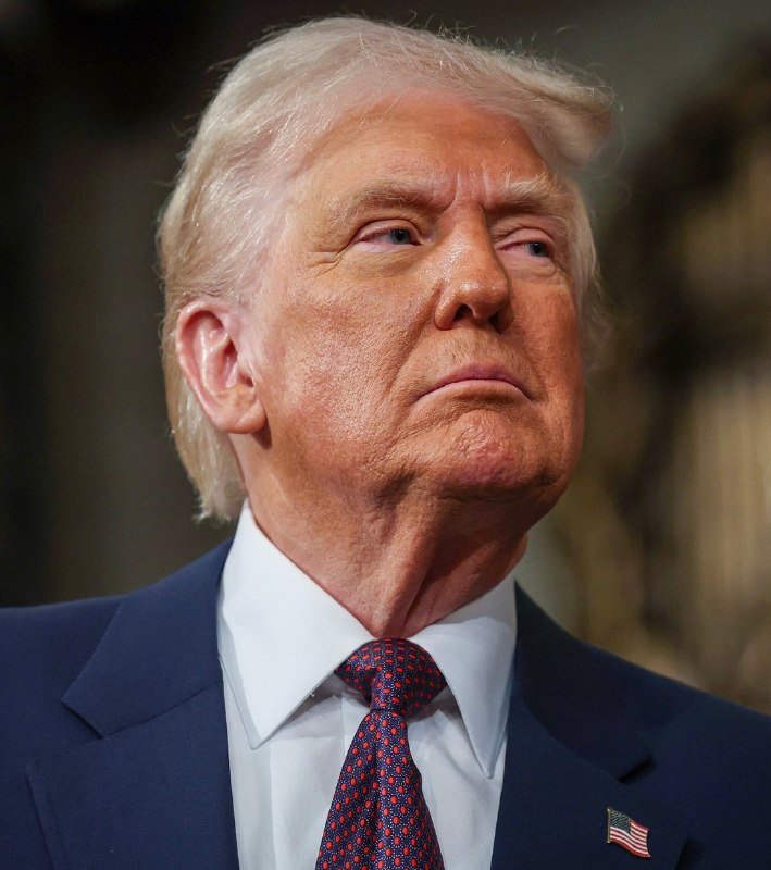 A man with blonde hair wearing a dark blue suit, white shirt, and red patterned tie with an American flag pin looks upward against a blurred background.