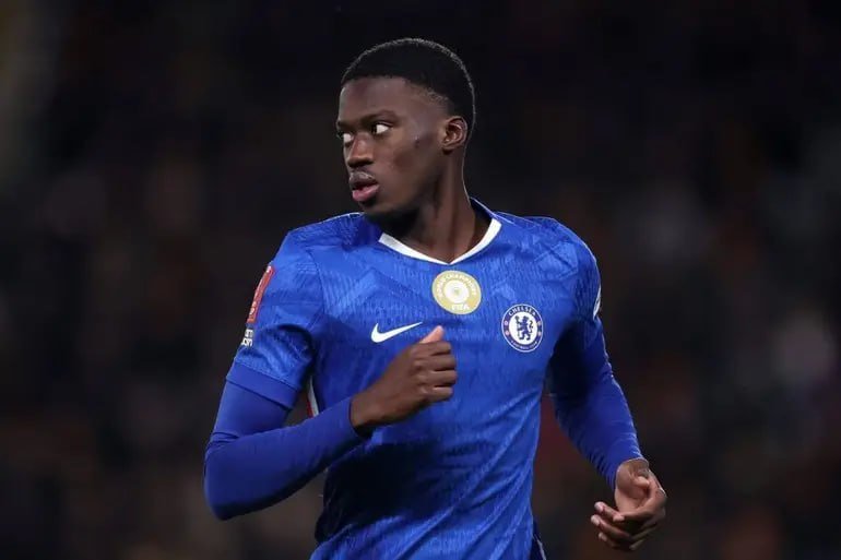 A soccer player in a blue Chelsea FC jersey runs on the pitch, looking to his right during a match.