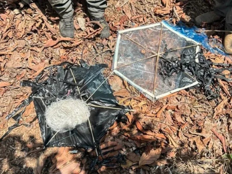 Two burnt, hexagonal kite frames—one with white string and black plastic covering, the other charred—lie on leaf-covered ground near a person's boots.