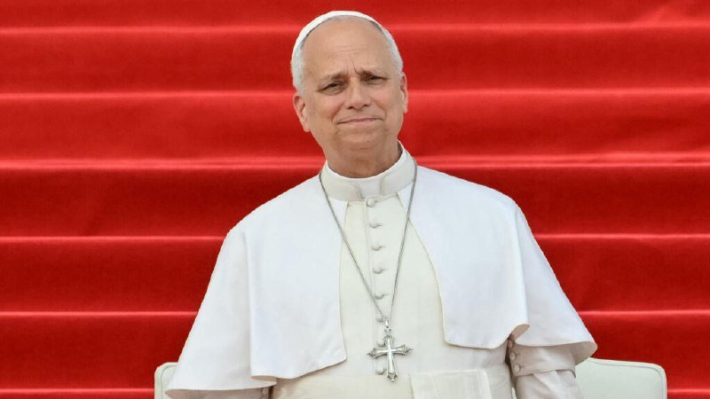 A man in white papal vestments and a zucchetto stands before a red-carpeted staircase, wearing a silver pectoral cross.