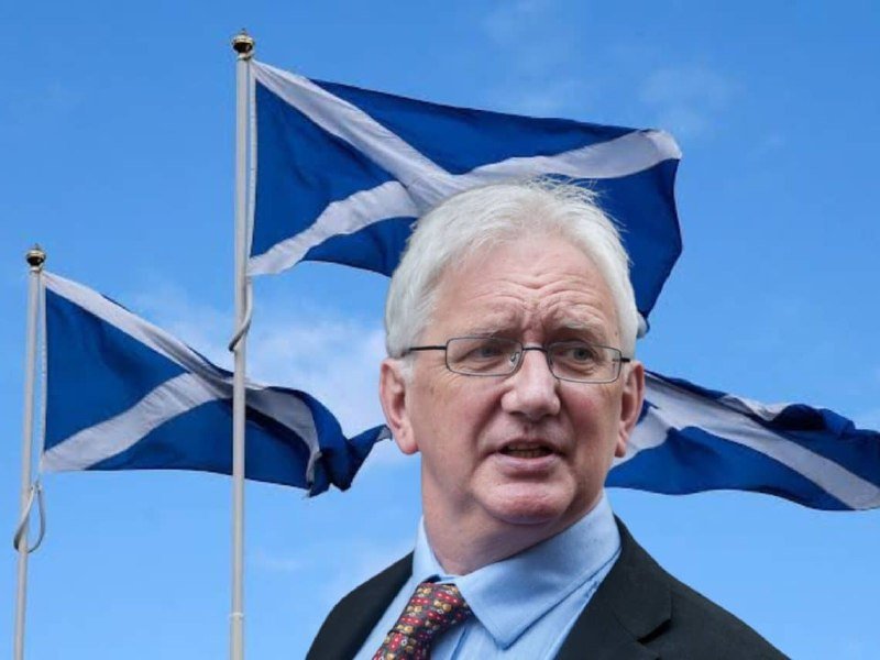 A white-haired man in glasses, a dark suit, and patterned tie speaks outdoors with two Scottish Saltire flags flying behind him against a blue sky.