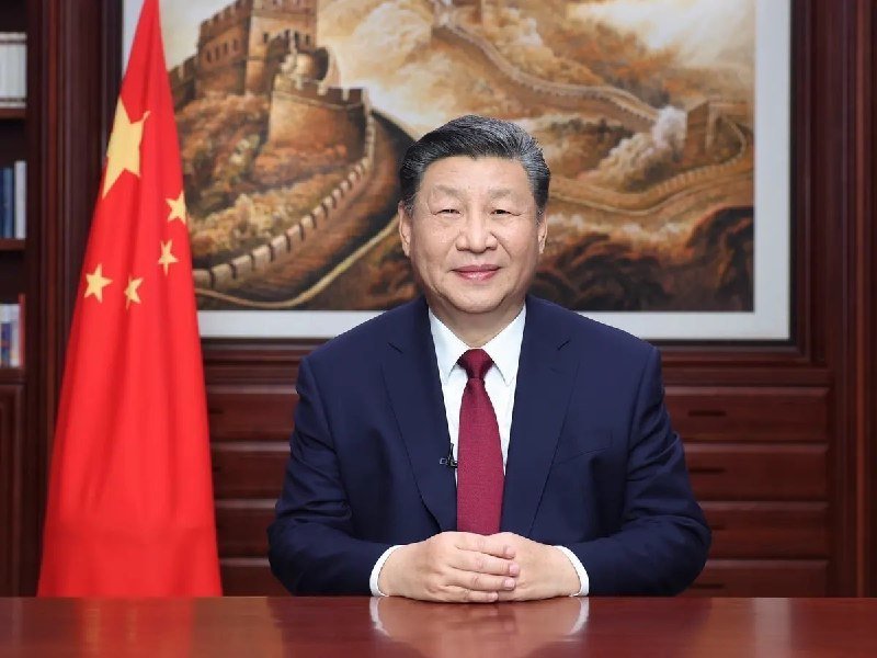 A man in a dark blue suit and red tie sits at a wooden desk, positioned before a Chinese national flag and a large painting of the Great Wall.