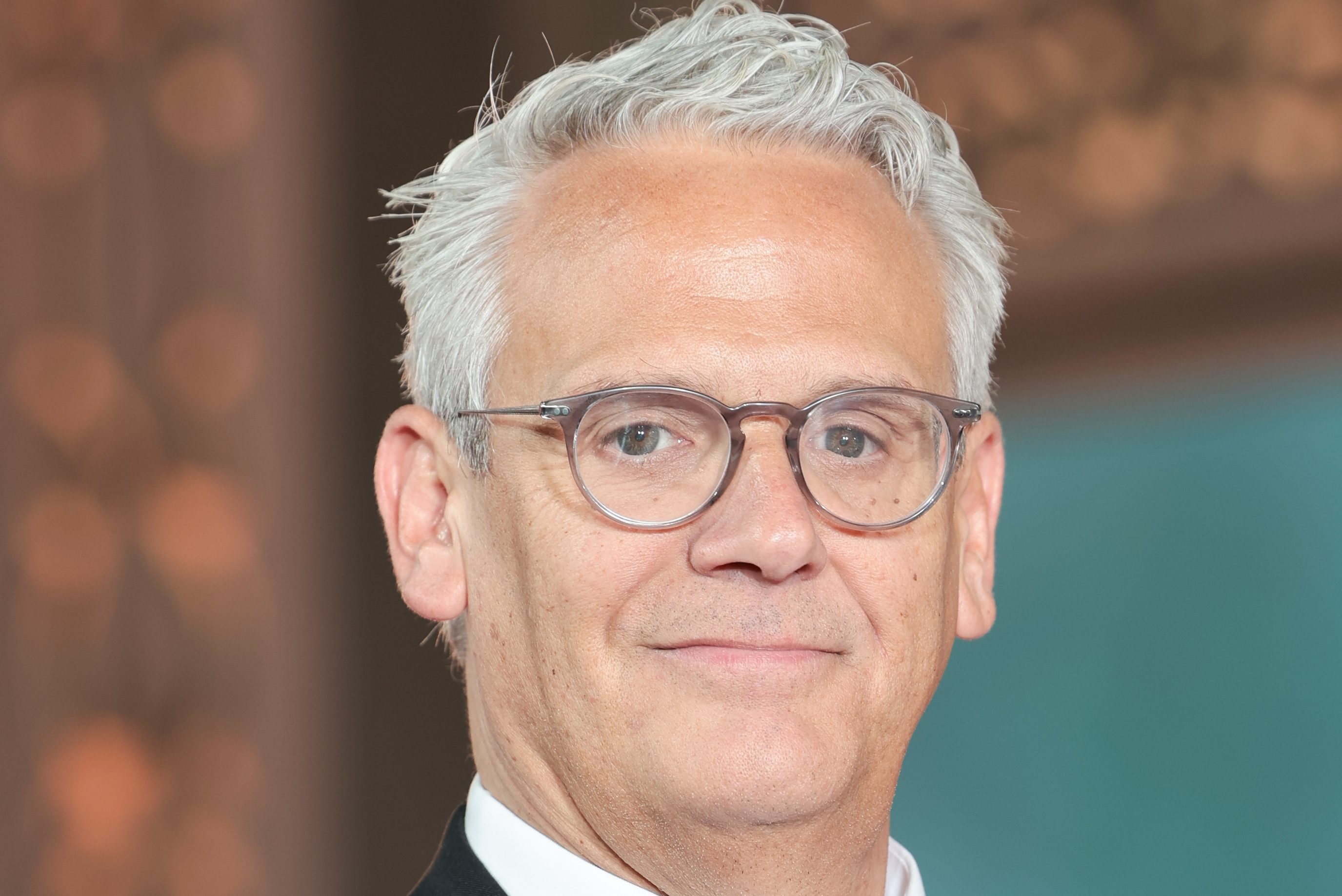 Close-up of a gray-haired man wearing round tortoiseshell glasses and a dark suit with a white shirt, posed against an orange and teal backdrop.