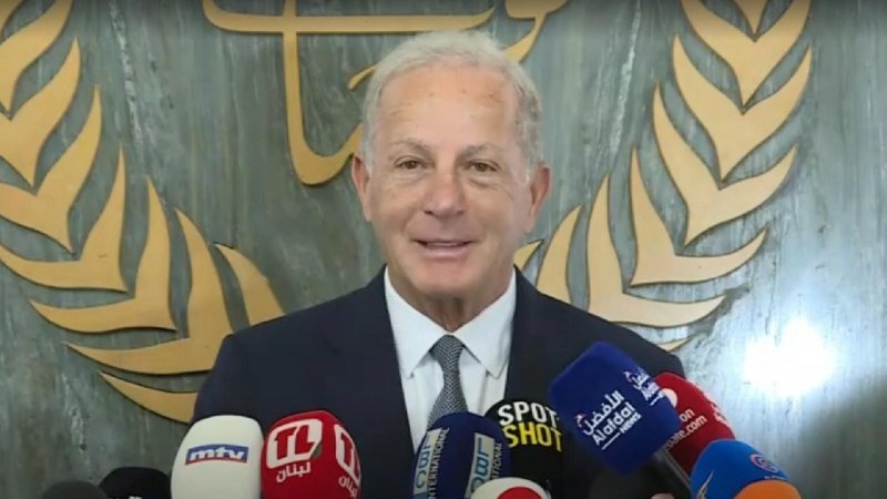 A gray-haired man in a dark suit speaks at a press conference, standing before microphones from MTV, OTV, and other outlets, with a decorative gold emblem behind him.