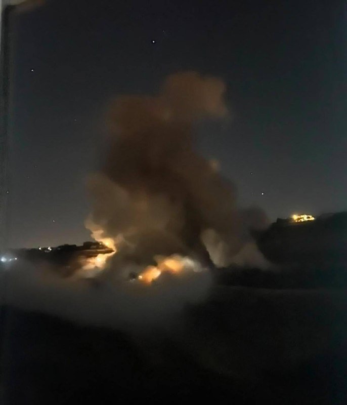 A large plume of smoke rises into a dark night sky above glowing fires burning at ground level, with distant lights visible on the horizon.