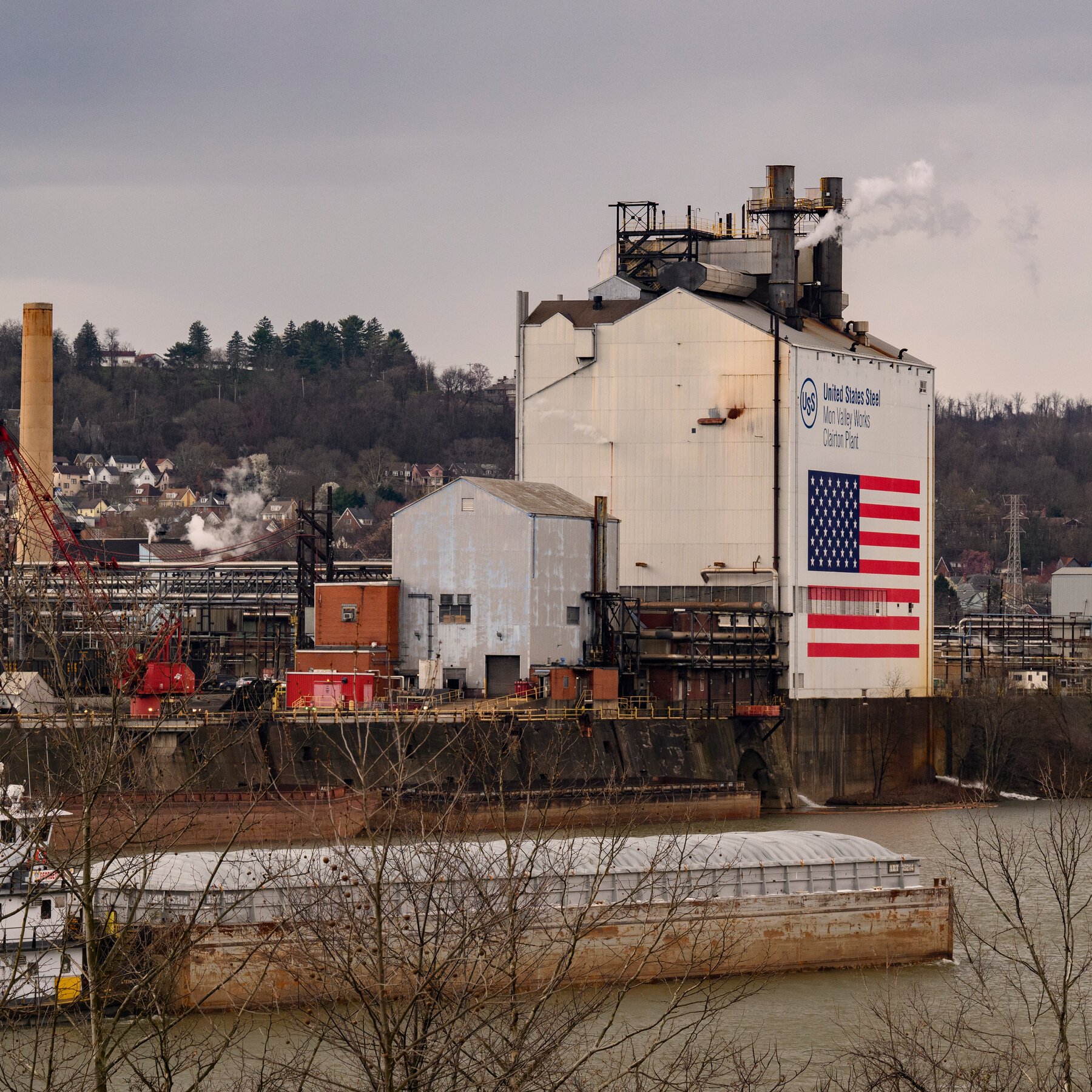 A riverside industrial facility displays a large "United States Steel, Mon Valley Works, Clairton Plant" sign and American flag mural on its white building, with smokestacks emitting steam above a covered barge on the water.