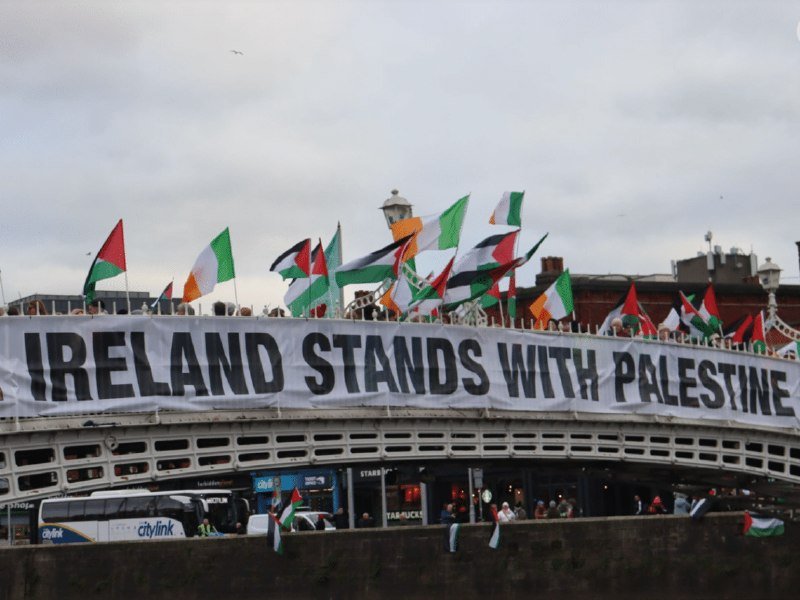 Demonstrators on a bridge wave Palestinian and Irish flags above a large banner reading "IRELAND STANDS WITH PALESTINE," with a "citylink" bus visible below.