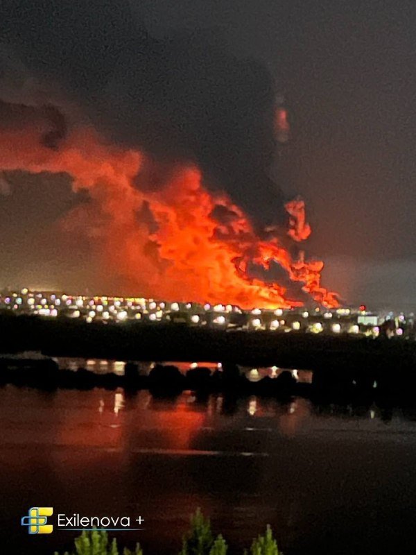 A large fire burns brightly at night above a cityscape, with city lights visible below and the flames reflecting on a body of water in the foreground.