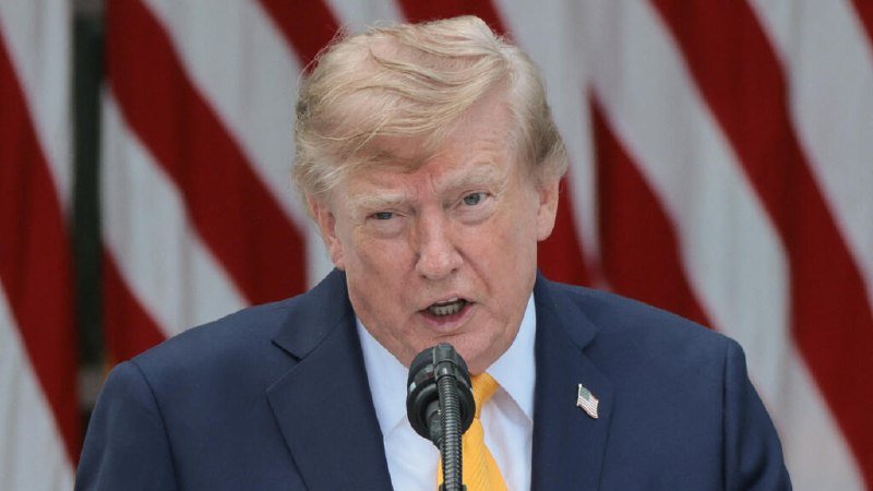 A blonde-haired man in a dark blue suit and yellow tie speaks into a microphone, with red and white striped fabric visible in the background.