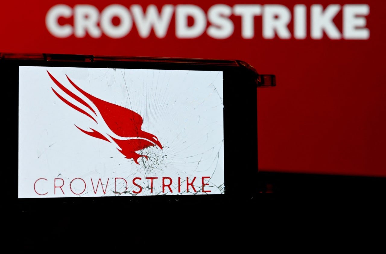 A smartphone displaying the cracked-screen CrowdStrike falcon logo is held against a red backdrop bearing the word "CROWDSTRIKE."