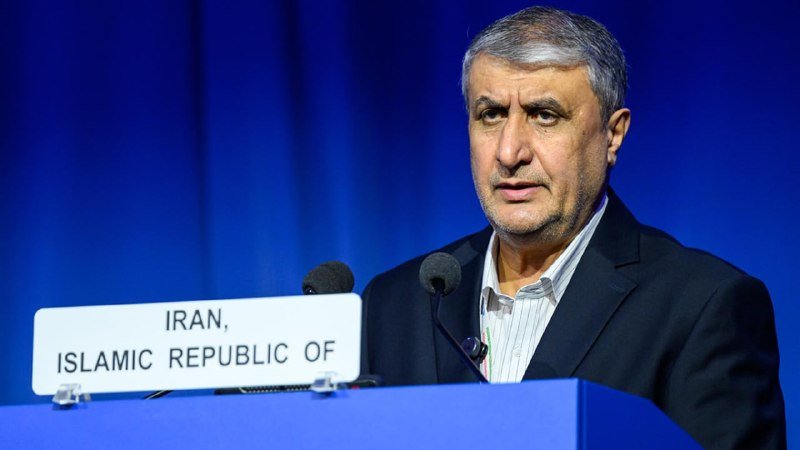 A gray-haired man in a dark suit speaks at a podium bearing a placard reading "IRAN, ISLAMIC REPUBLIC OF" against a blue backdrop.