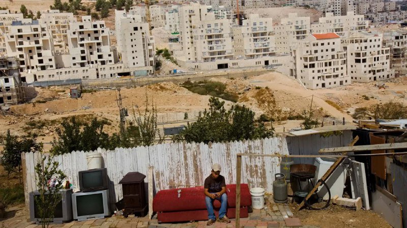 A man sits on a red couch outdoors behind a weathered white fence, with discarded televisions, a gas cylinder, and a bicycle nearby, overlooking a hillside of densely packed unfinished concrete buildings.