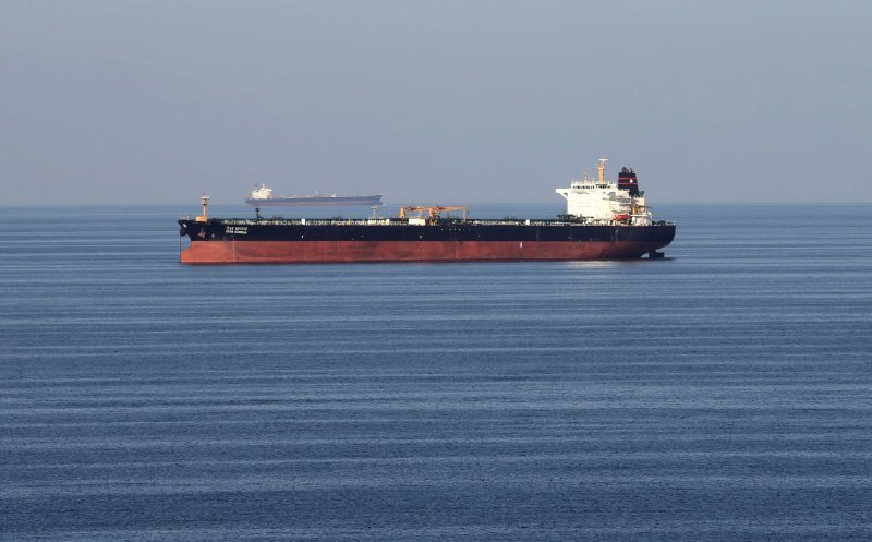A large red and black tanker ship floats on calm blue waters, with a smaller cargo vessel visible in the hazy distance behind it.