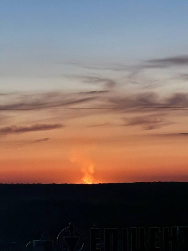 The sun sets below a dark horizon beneath an orange and blue sky streaked with wispy clouds, as a power line crosses the frame above a silhouetted watermark.