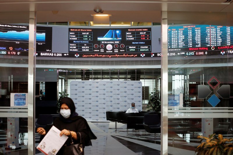A woman in a black abaya and face mask walks past electronic stock tickers displaying the Bahrain All Share Index, with a seated man visible through a glass entrance behind her.