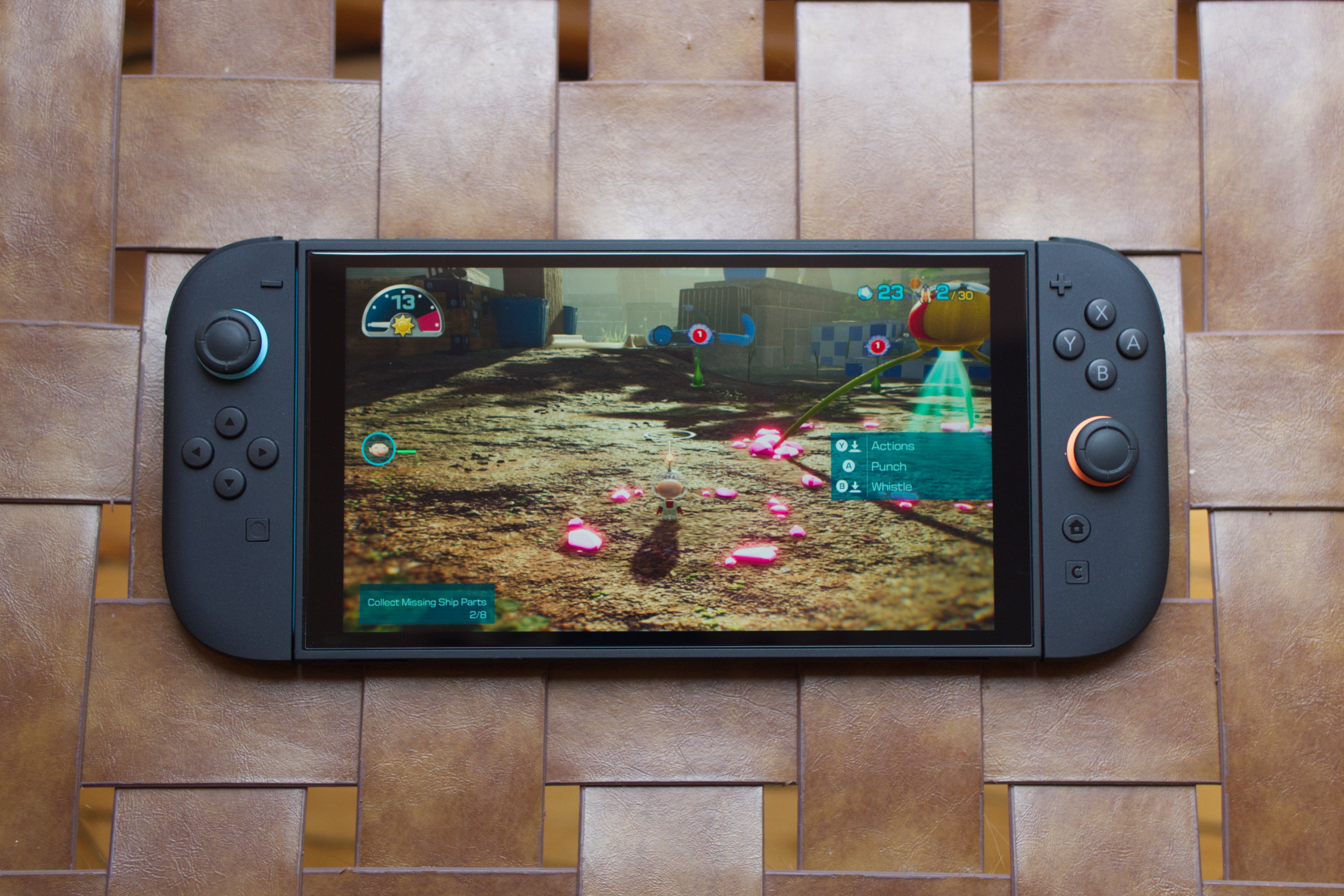 A handheld gaming console displays a video game featuring a small character collecting pink items on a textured surface, resting on a brown woven mat.