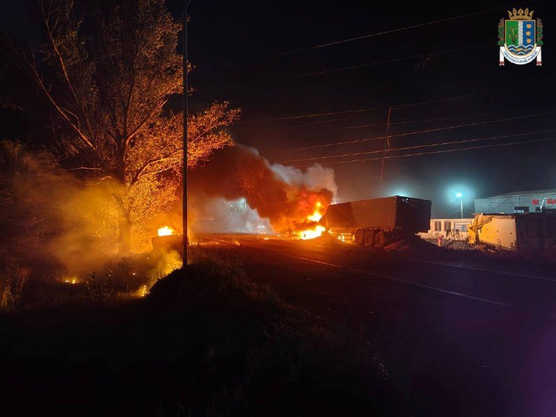 A truck and surrounding vegetation burn with large flames and smoke at night along a roadside, with an emblem visible in the corner.